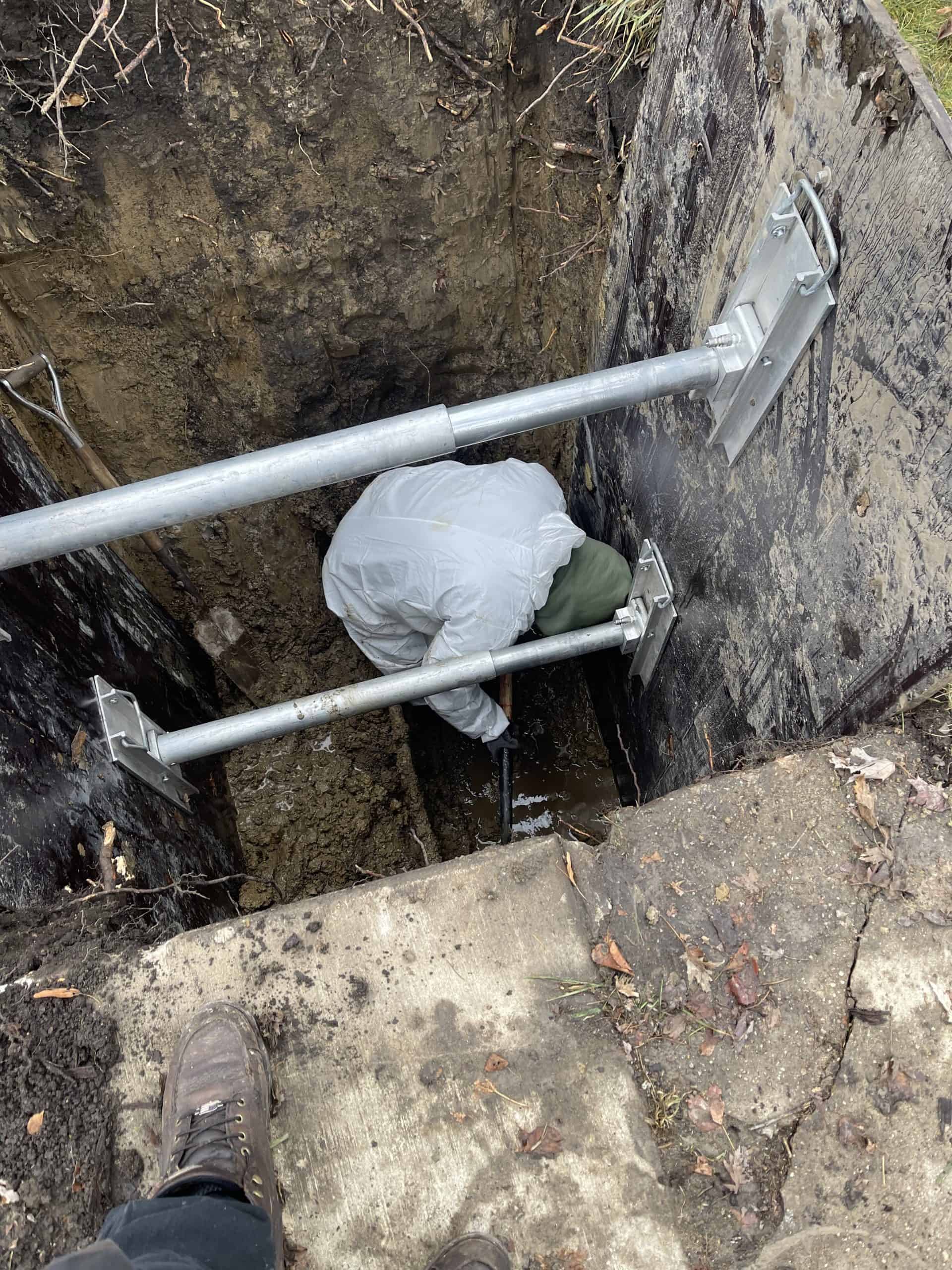 A person in a white protective suit works diligently in a deep, narrow trench reinforced with metal supports. The trench has wooden walls and is surrounded by concrete and soil, while a pair of boots stands at the edge—a typical scene during hydro jetting services in Chicago, IL.