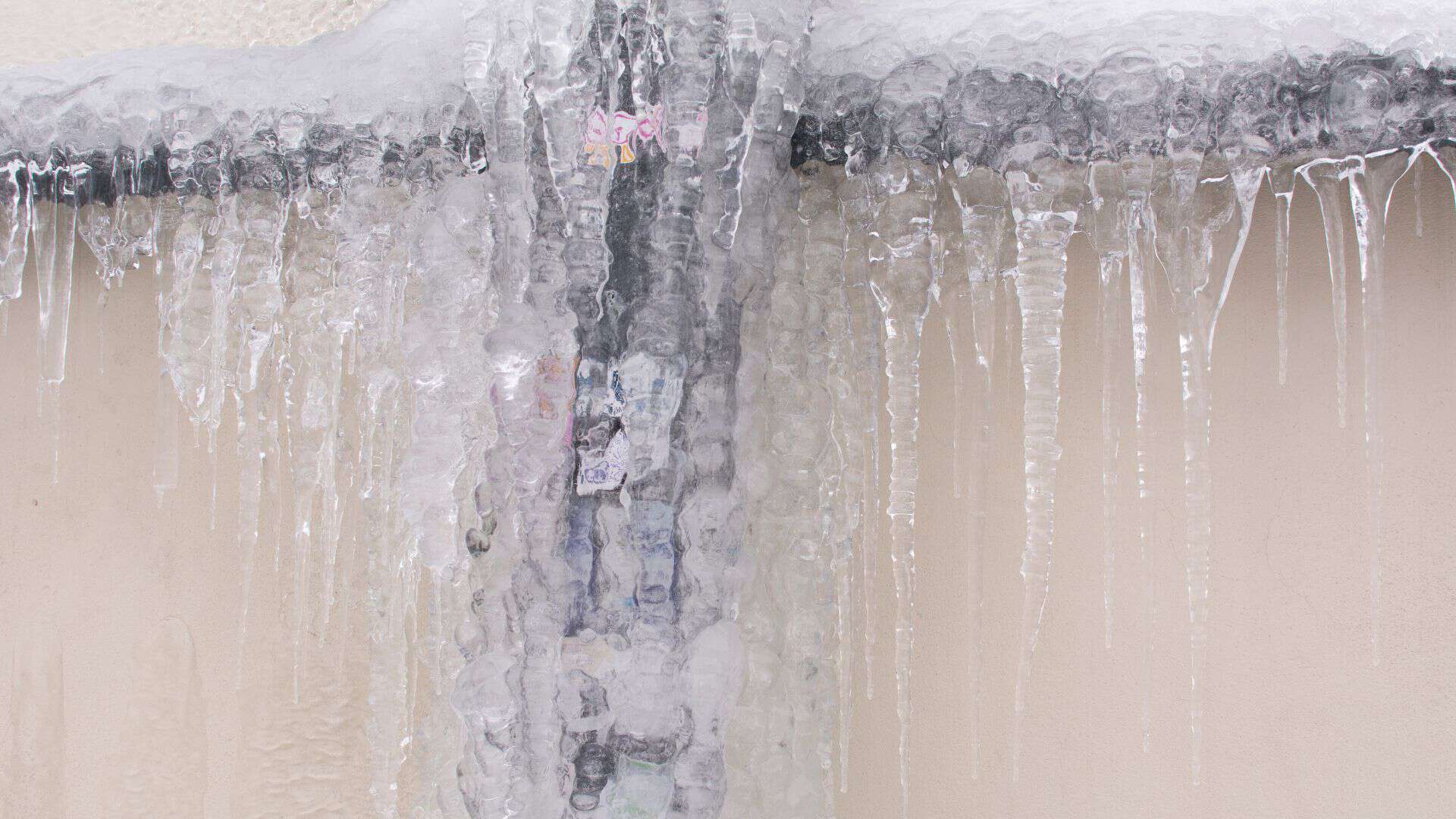 Icicles hang from a frozen pipe against a beige wall. The ice formation surrounds the pipe, with long icicles dangling down, creating a cold and wintry scene. For such frigid plumbing emergencies, trust the best Chicago plumbing company to keep things flowing smoothly.
