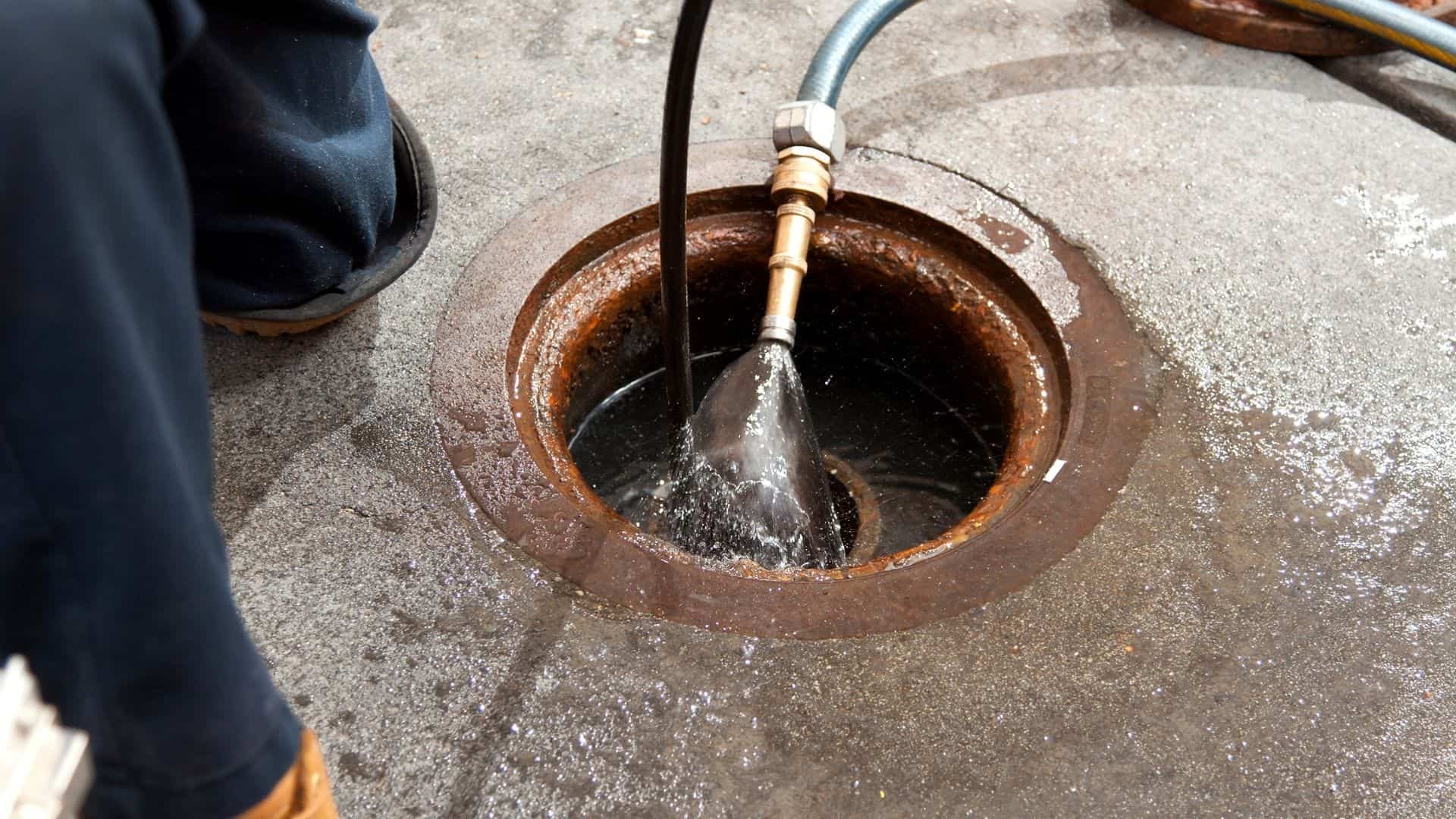A person uses a hose for hydro jetting in Chicago, cleaning a large circular manhole on a concrete surface. Water sprays from the hose, creating splashes around the rim. Nearby, part of a worker's boot is visible.