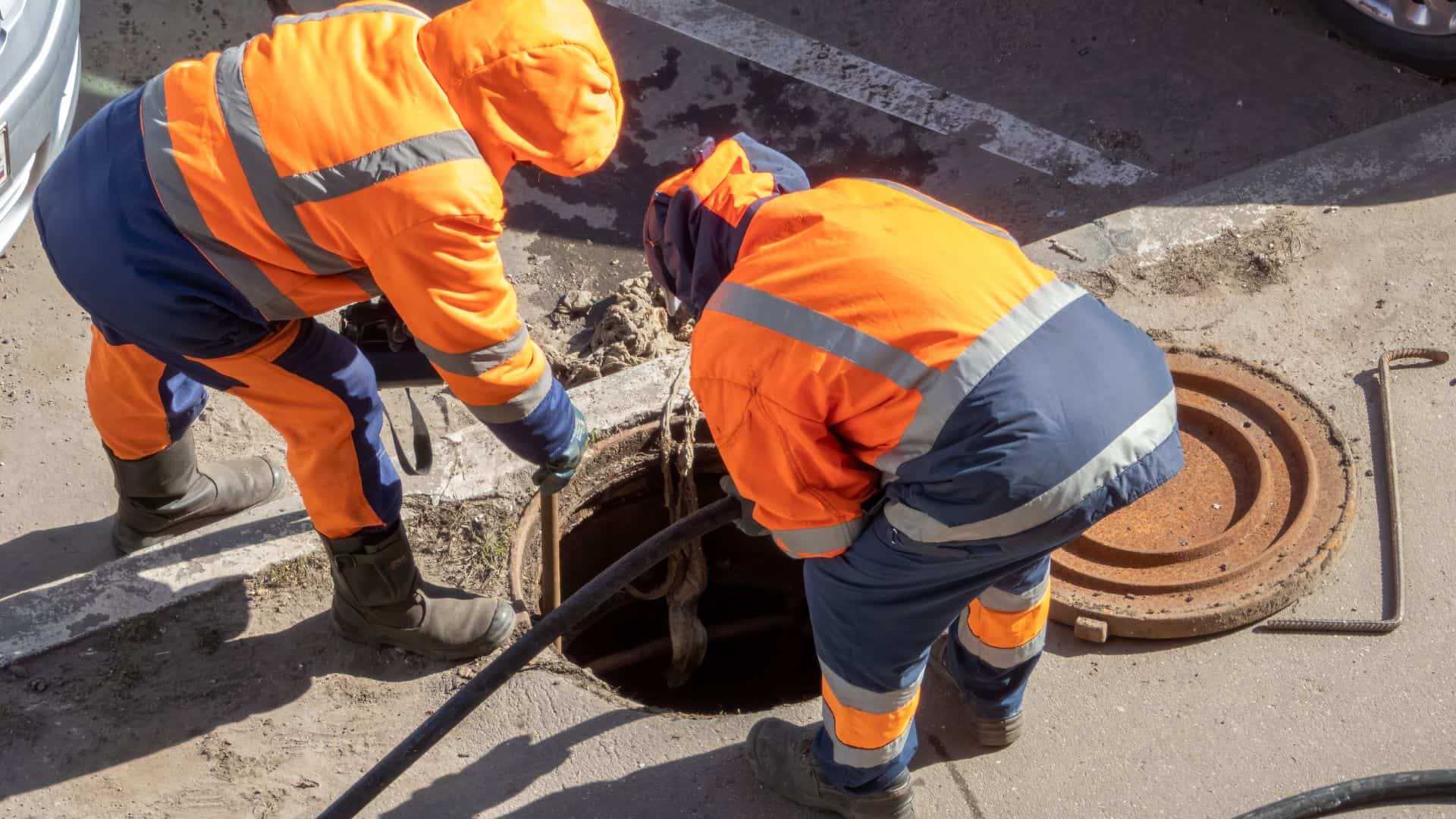 Two workers in orange high-visibility clothing are inspecting a manhole. They are bending over, holding a hose or cable, with the manhole cover nearby.