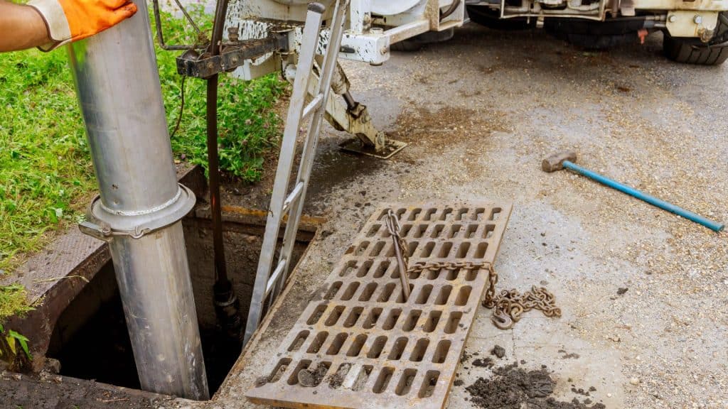 A person wearing an orange glove operates a large metal pipe into a street drain, reflecting the expertise of the best Chicago plumbing company. A ladder and open grate are nearby, alongside a small chain and hammer. The scene is by a grassy area with a partially visible vehicle.