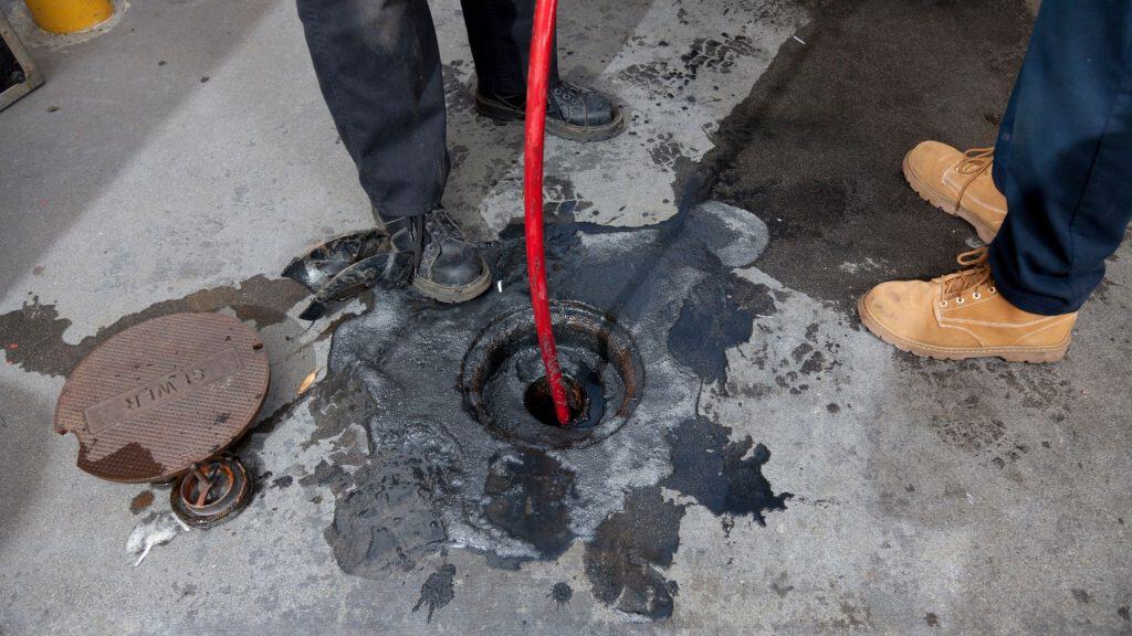 Two people use a red tool to clean a drain on a concrete surface.