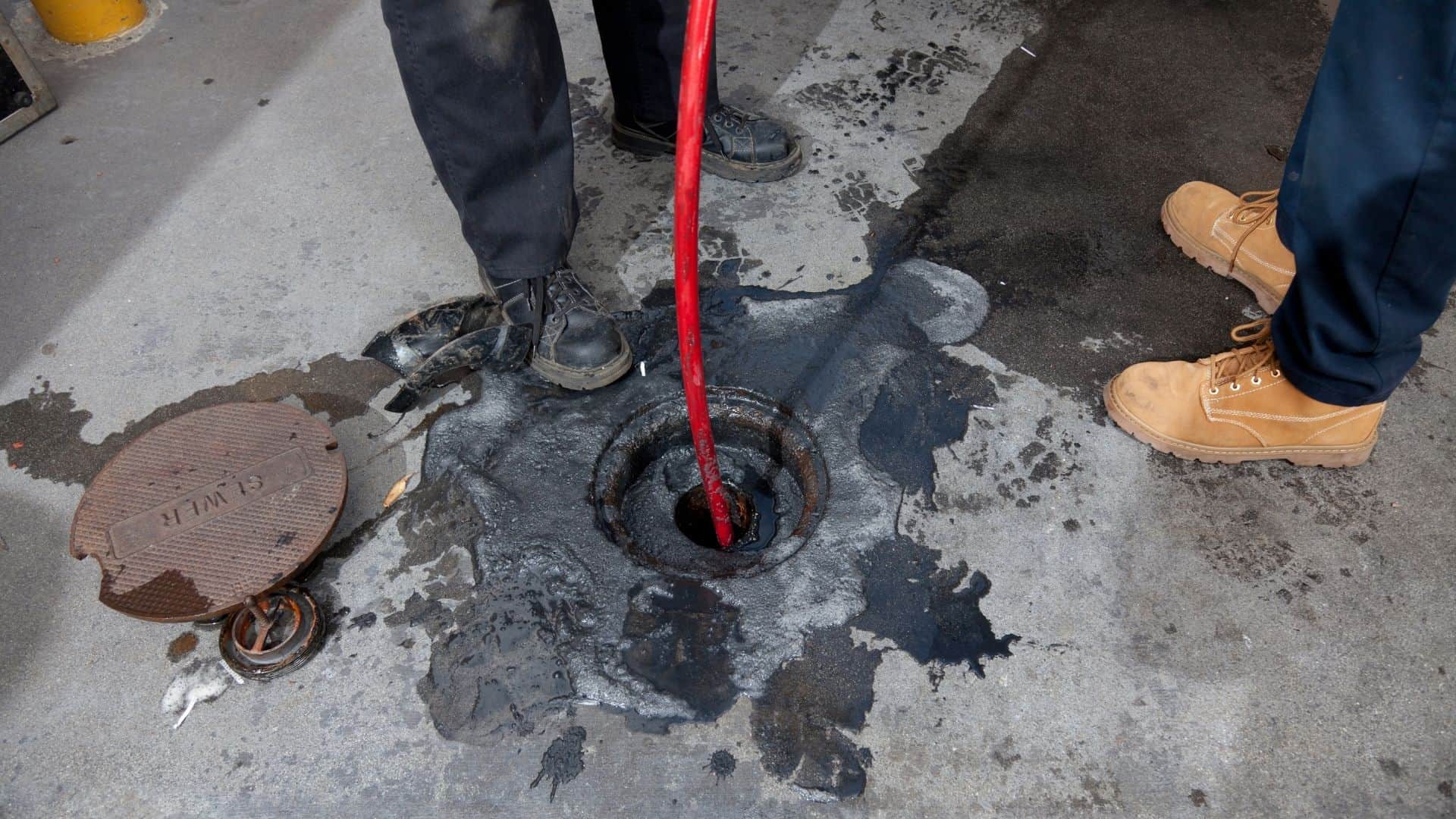 Two people are using a red tool to clean a drain on the concrete surface, employing techniques popular with the best Chicago plumbing companies. Wet grime surrounds the area, and nearby are a manhole cover and a small drain cover. One wears black shoes, the other brown boots.