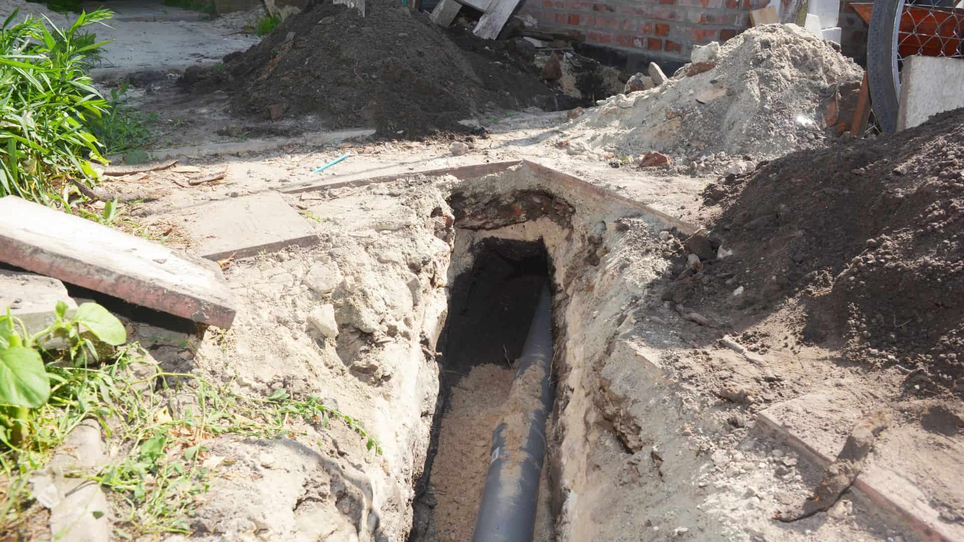 A trench with an exposed underground pipe is surrounded by piles of dirt and construction materials. The area appears to be a work site, likely managed by the best Chicago plumbing company, with greenery on one side and a partially visible brick structure in the background.