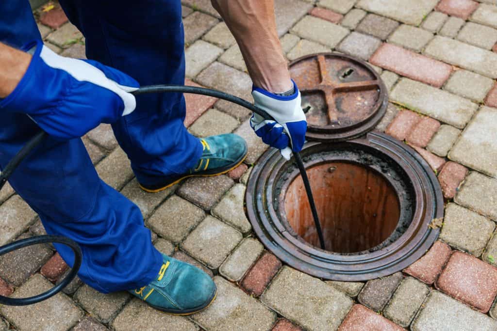 A worker in blue protective gloves and overalls from the best Chicago plumbing company uses a long tool to unclog a round outdoor drain on a paved surface. The drain cover is partially removed, and the pavement consists of multicolored bricks, showcasing top-notch sewer repair in Chicago.