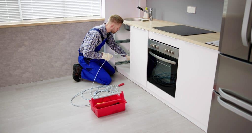 A man in blue overalls is kneeling and installing a pipe under a kitchen cabinet. There is a red toolbox on the floor next to him, representing the best Chicago plumbing company. The kitchen features white cabinets, a stainless steel oven, and a grey backsplash.