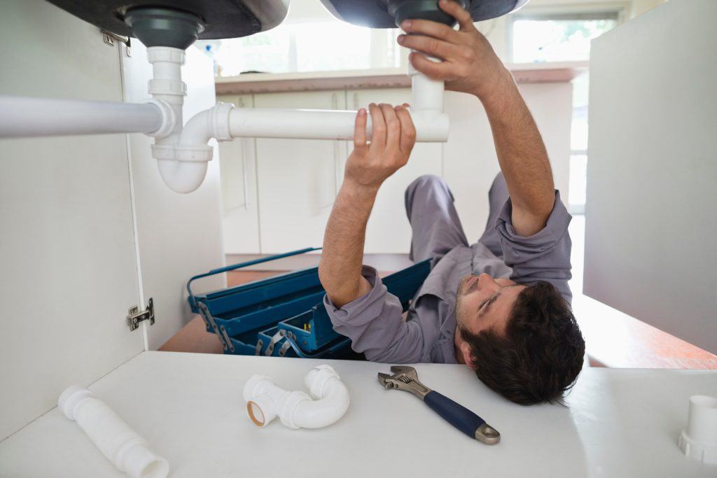 A person wearing a gray uniform is lying on their back under a sink, fixing pipes. Various plumbing tools and pipe fittings are scattered around, including a wrench. A toolbox is open beside them.