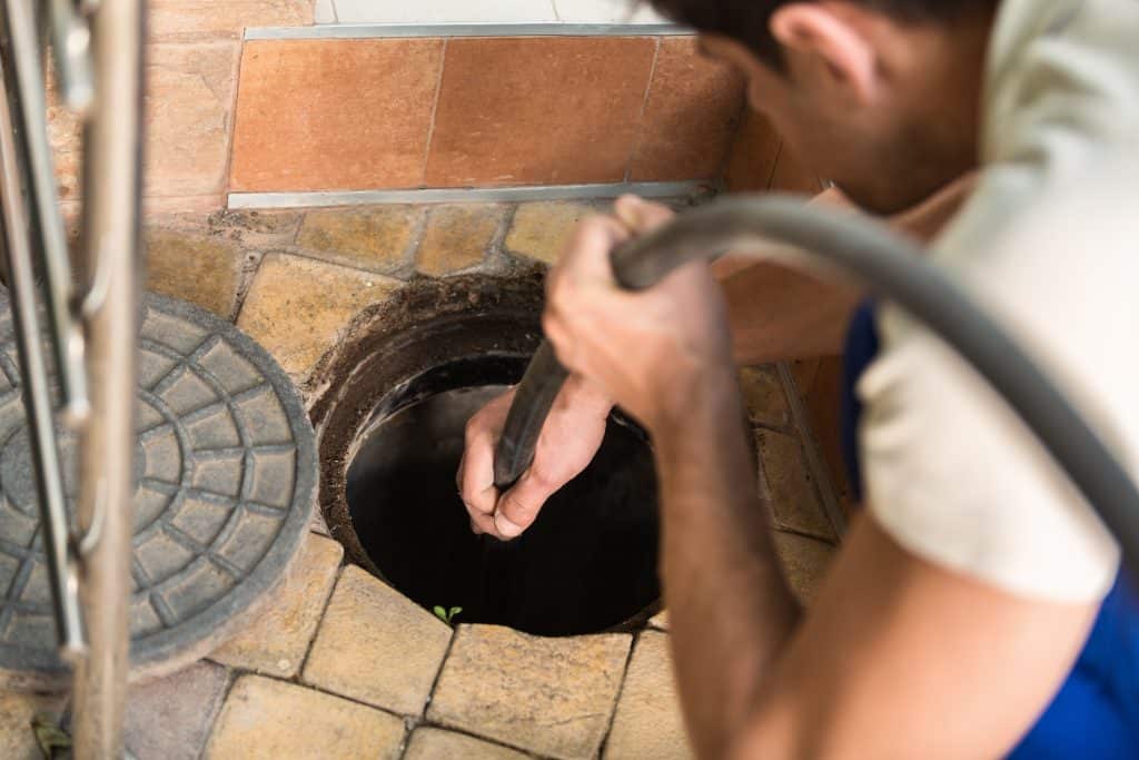 A person wearing a blue apron is focused on inspecting a drain with a hose, showcasing expert plumber skills in Chicago. The scene unfolds on a tiled floor, where meticulous hydro jetting takes place near the round drain cover.