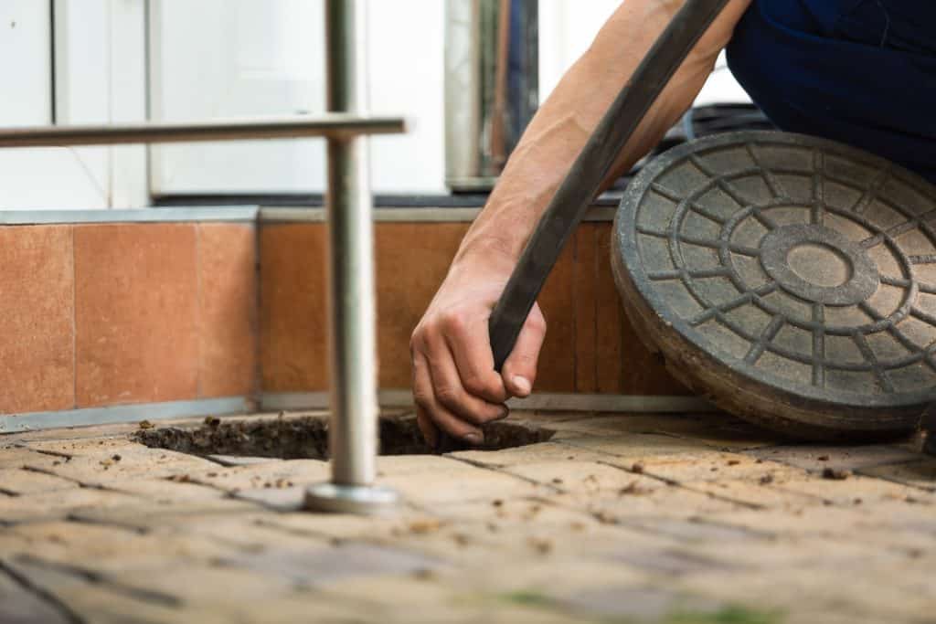 A person from the best Chicago plumbing company expertly lifts a sewer manhole cover with a metal tool on a tiled surface. The scene captures a close-up of the hand and tool in action, with part of a railing visible nearby, illustrating their efficient work.