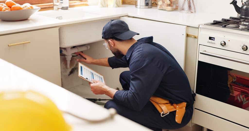 A plumber in a navy uniform and cap is inspecting pipes under a kitchen sink. He holds a tablet in one hand and is using a flashlight with the other. A tool belt is visible, and the kitchen counter has some oranges in a bowl.