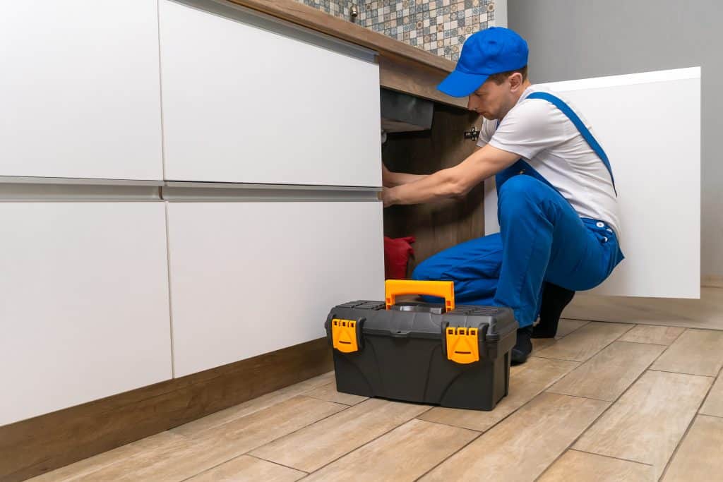 A plumber wearing blue overalls and a cap is crouched down, working under a kitchen sink. A black toolbox with yellow latches is on the tiled floor nearby.