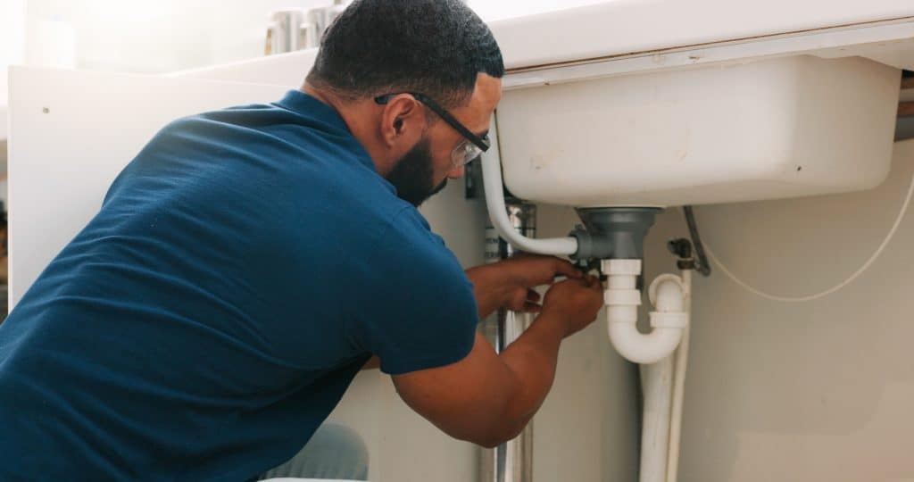 A person wearing a blue shirt is crouched down under a sink, using tools to fix or adjust the plumbing pipes. The focus is on the pipes, and it appears to be a kitchen or bathroom setting.