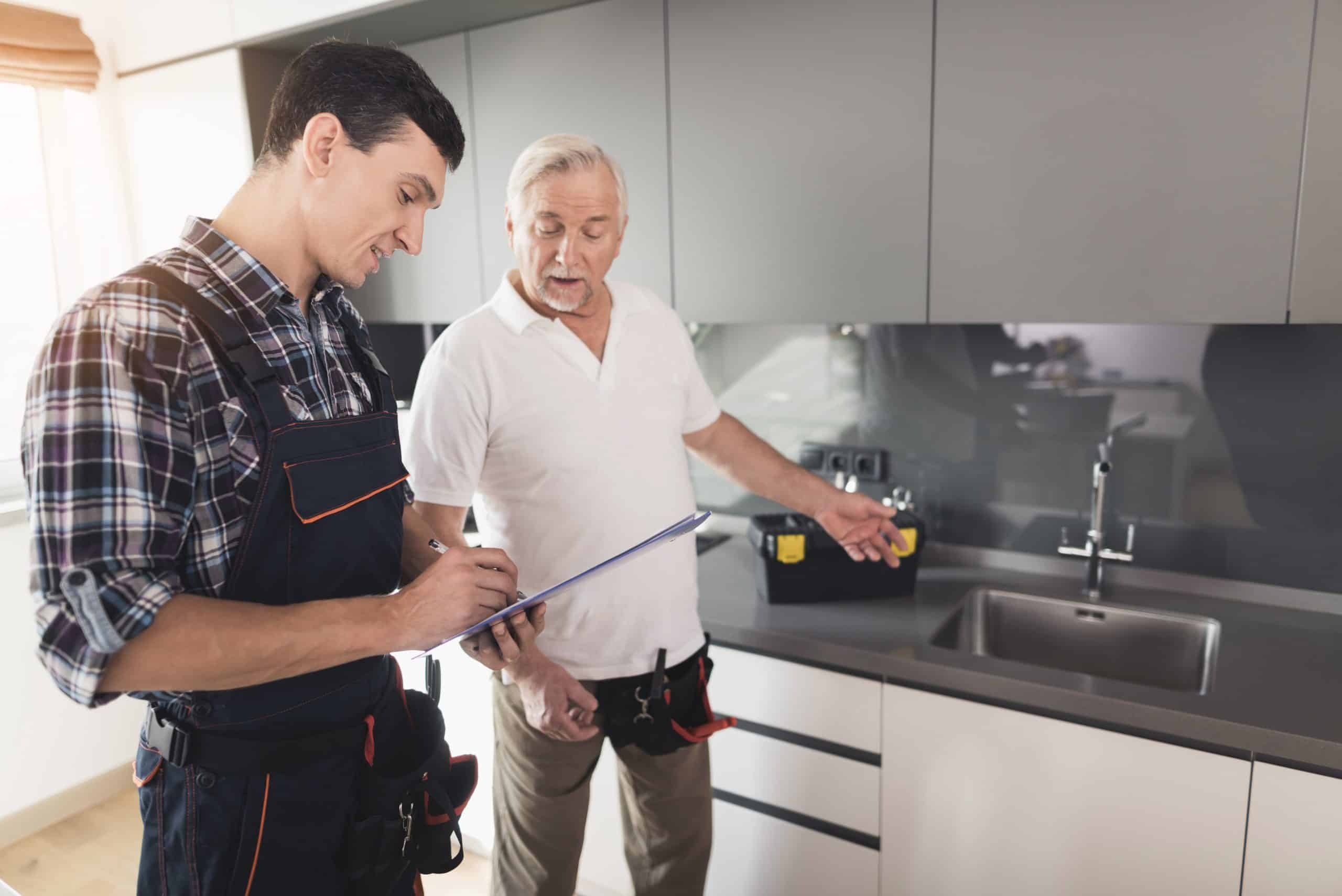 A young man in a checkered shirt and overalls writes on a clipboard while an older man in a white polo shirt gestures toward a kitchen sink. They appear to be discussing a plumbing or home repair task.