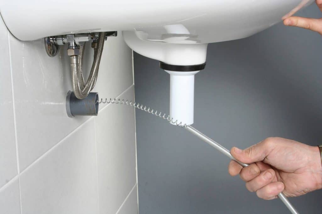 A person uses a plumber’s snake to unclog the drainpipe under a white sink mounted on a tiled wall in Cook County, Illinois