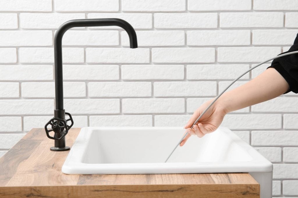 A person holds a plumbing snake inside a white sink with a black faucet, set against a white brick wall background in Cook County, IL