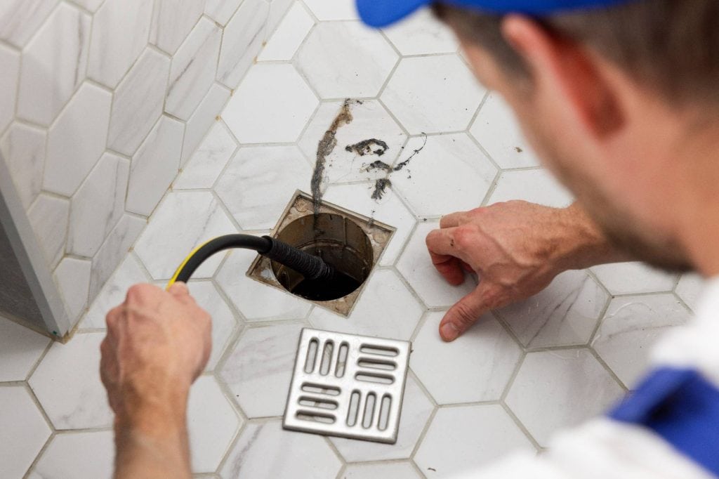 A plumber uses a flexible tool to unclog a floor drain in a tiled bathroom, with the drain cover removed and placed nearby.