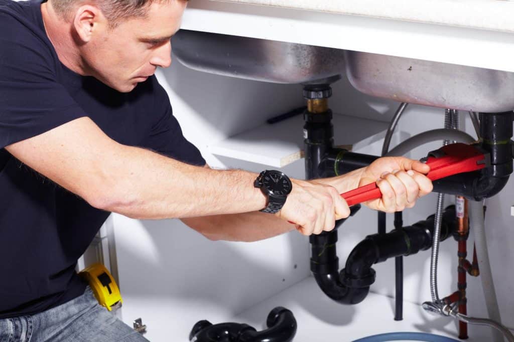 A plumber is using a red wrench to tighten or loosen a pipe joint under a kitchen sink, surrounded by various plumbing fixtures and pipes.