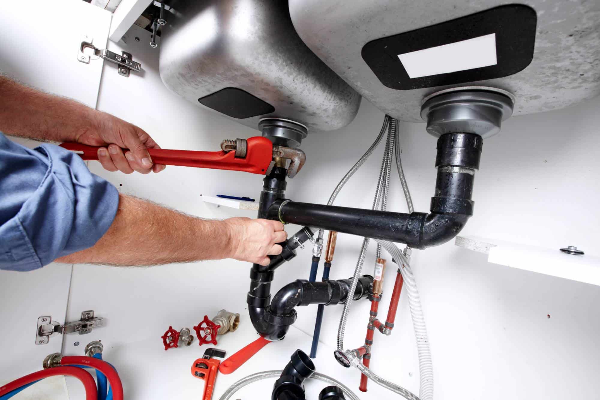 A plumber uses a red pipe wrench to tighten a black pipe under a kitchen sink, surrounded by various pipes, fittings, and plumbing tools inside a cabinet.