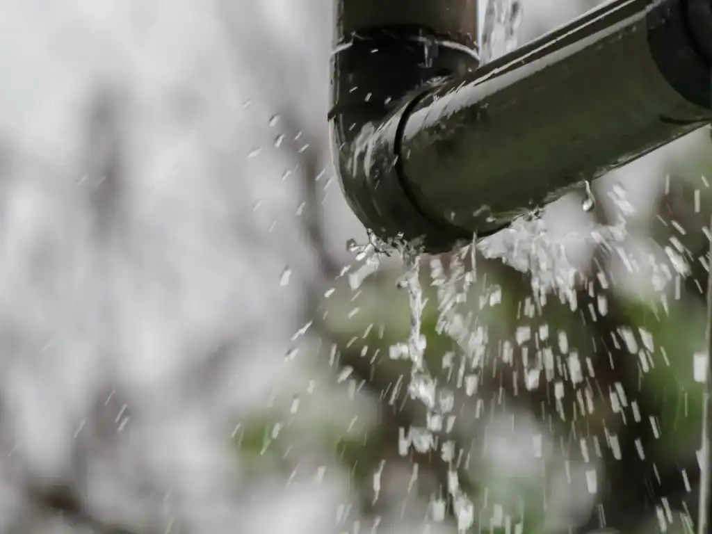 Close-up of rainwater pouring from the end of a metal gutter, with water droplets splashing and falling—a reminder to call a plumber in Chicago, IL if your gutters or drainage need expert attention. Blurred tree branches and cloudy sky in the background.