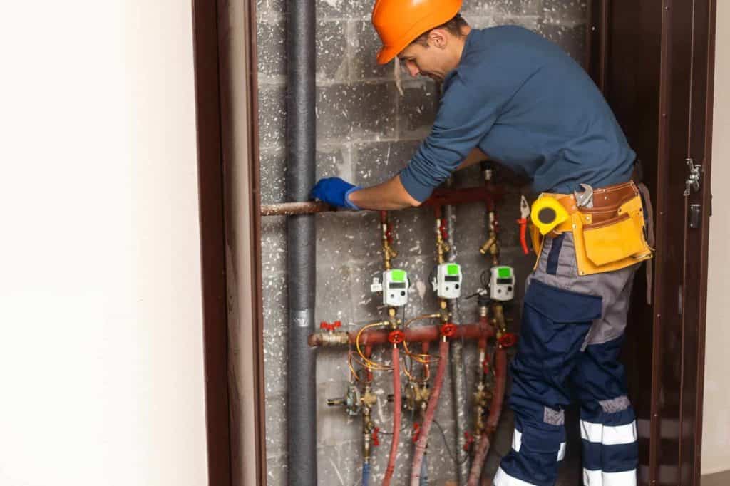 A plumber Chicago, IL, wearing a hard hat, gloves, and a tool belt works on pipes and valves inside a utility closet with exposed brick and multiple water meters.