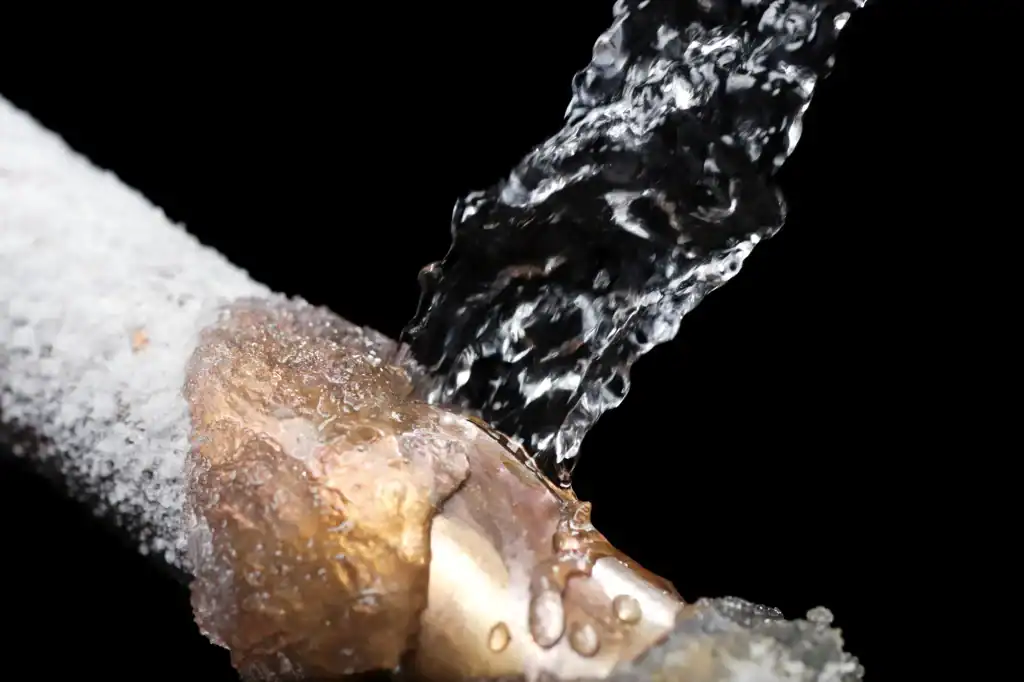 A close-up of a frozen and burst pipe with water gushing out, set against a black background. The metal pipe is covered in ice, highlighting the damage caused by freezing temperatures.
