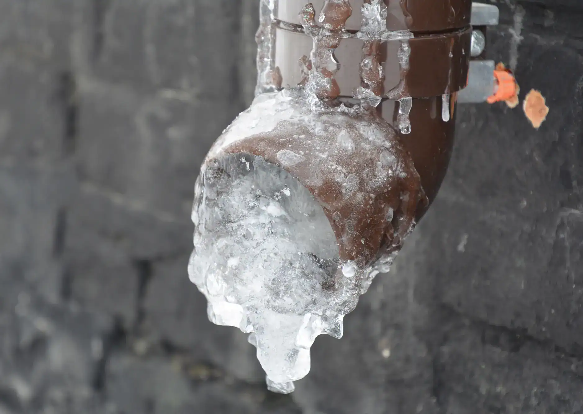 A brown outdoor pipe is covered in thick, clear ice, with icicles hanging from it against a dark brick wall, indicating freezing weather conditions.