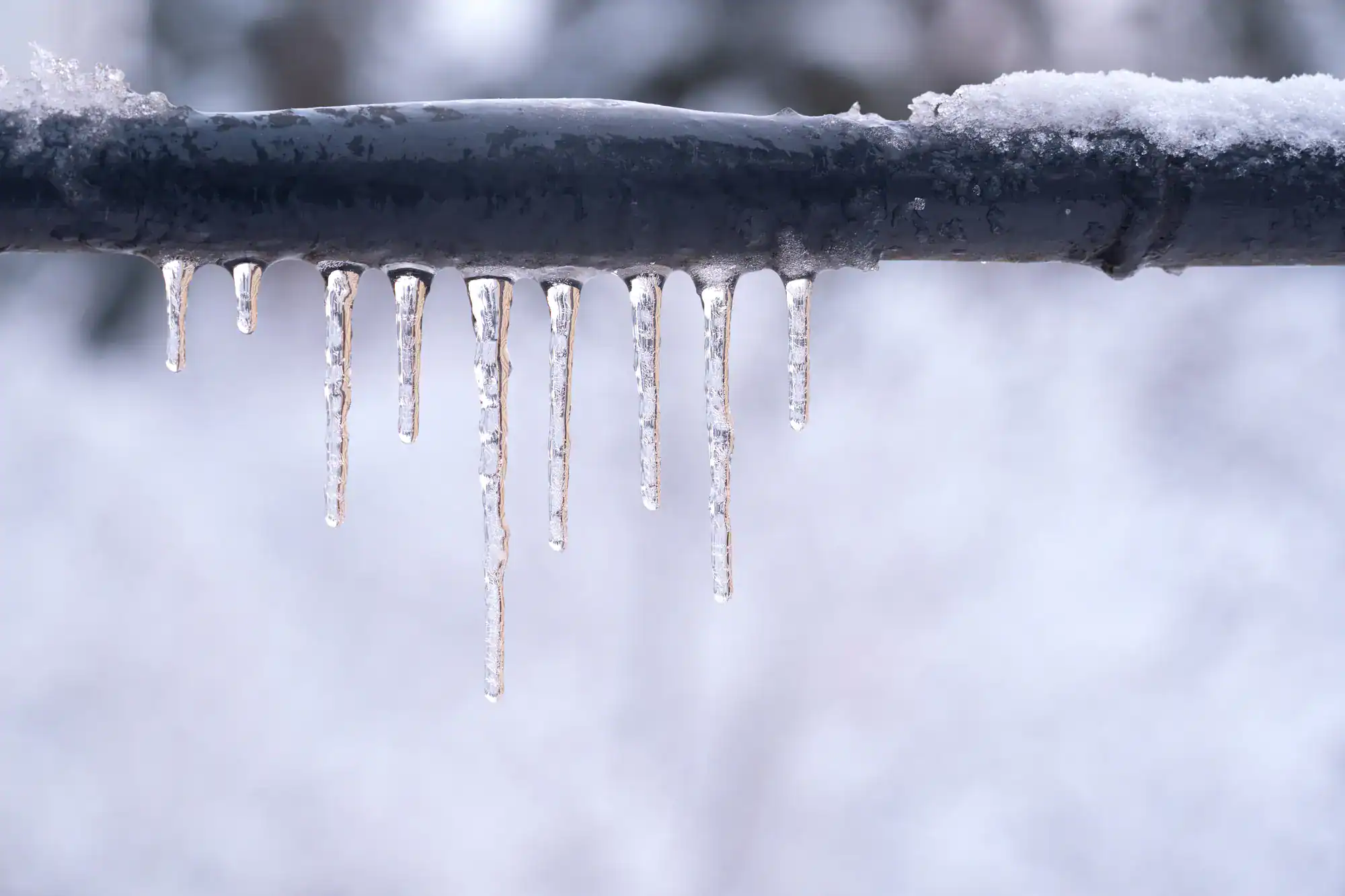 A row of icicles hanging from a snow-covered metal pipe, with a blurred snowy background.