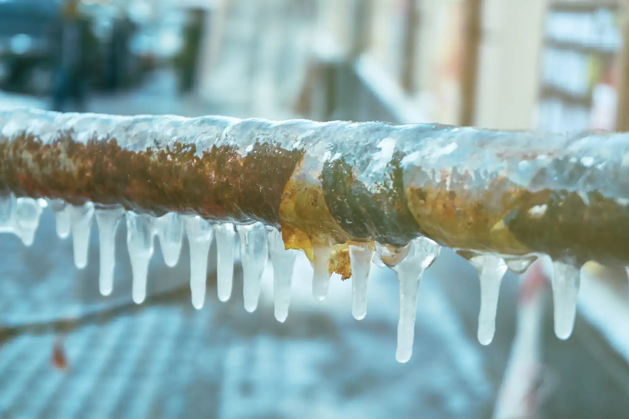 A rusty metal pipe covered in a layer of ice with long icicles hanging from it, set against a blurred urban background.