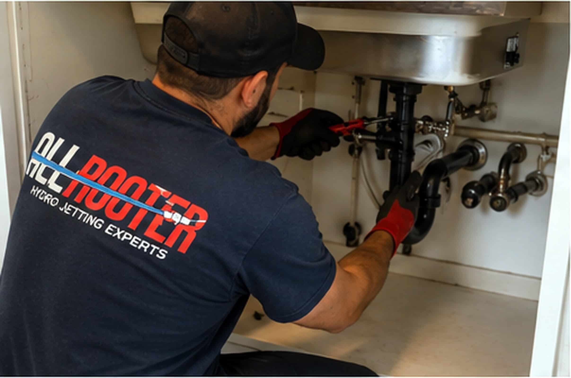 A plumber wearing gloves uses pliers to fix pipes under a sink, shirt reads “All Rooter Hydro-Jetting Experts.”